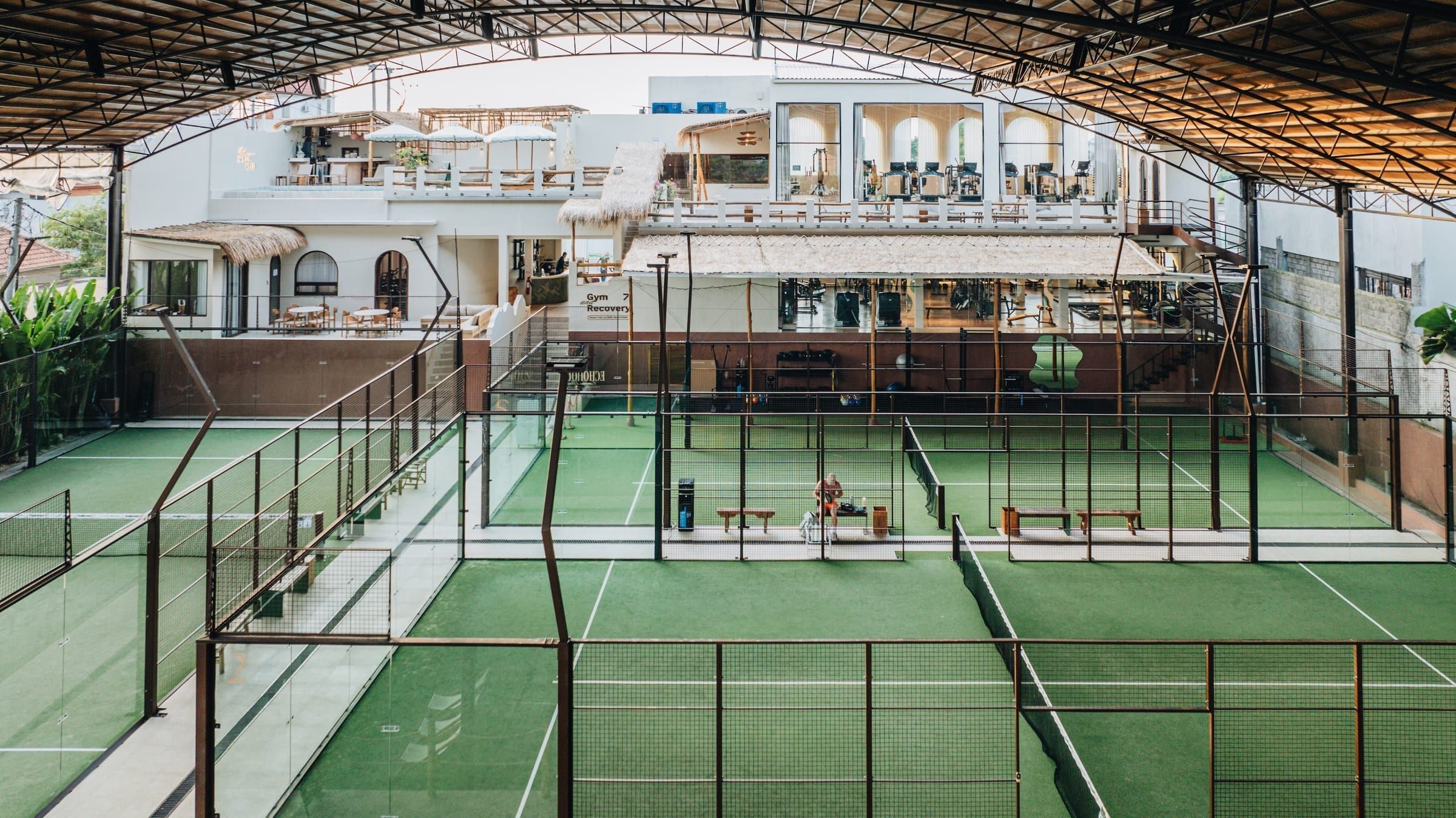 Padel courts under a curved roof, with the white clubhouse behind, at Bali Social Club