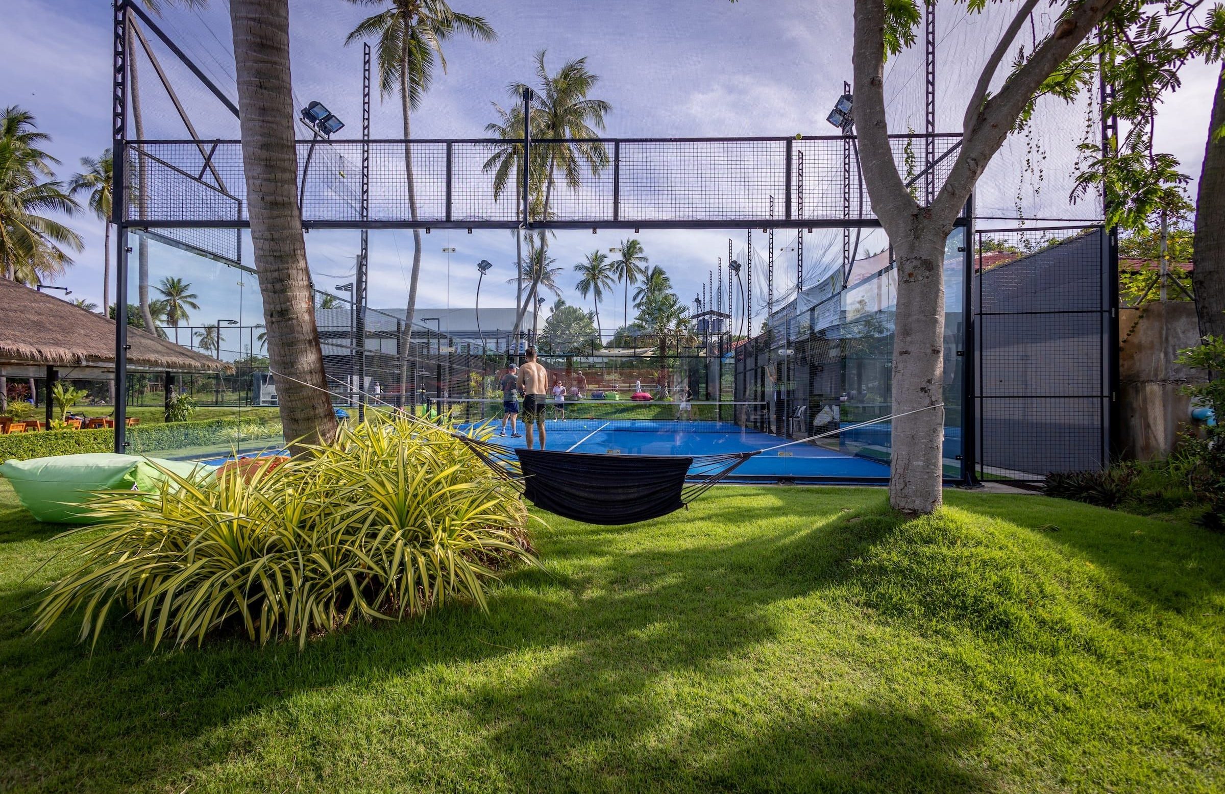 Blue padel court among palm trees, with a hammock on the lawn in front