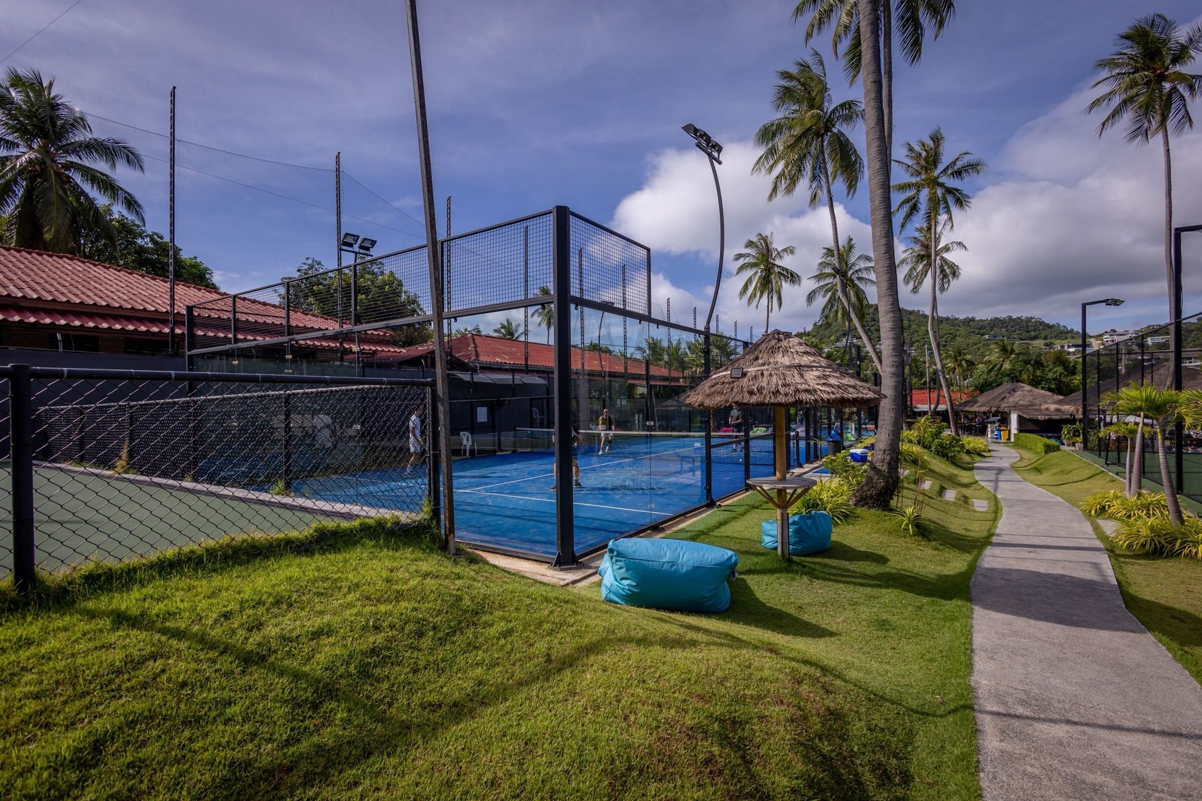 Path past a thatched shelter and beanbags beside the blue courts